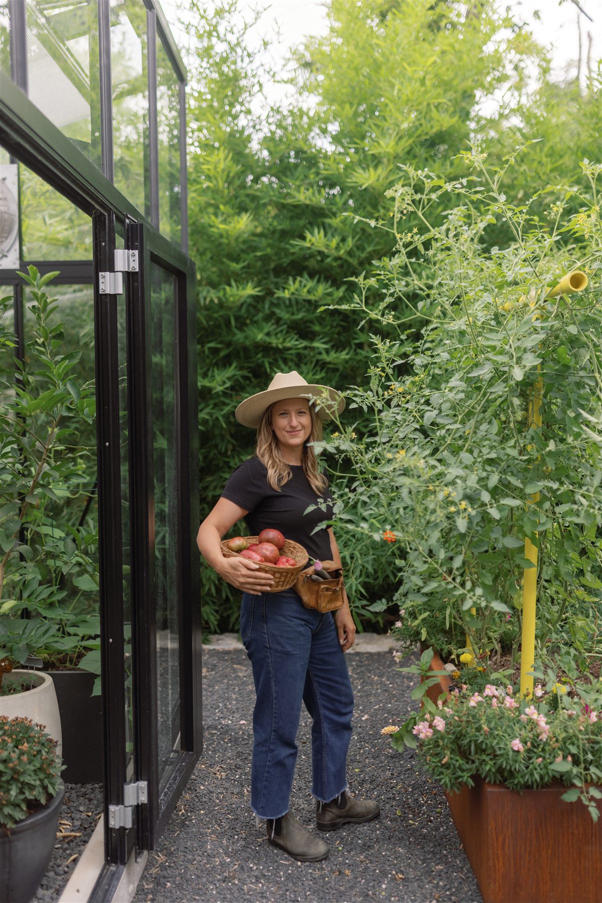 Kenley holding a basket of tomatoes beside a glass greenhouse and thriving garden
